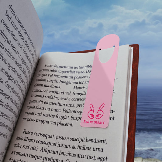 A pink paper book bookmark shaped like a bunny with the text 'BOOK BUNNY' printed on it, shown resting on an open book with a blurred beach background.