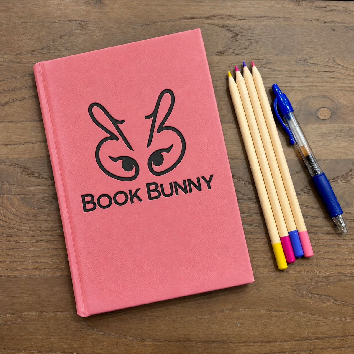 A pink hardcover notebook with a black bunny face logo and the text 'BOOK BUNNY' on the cover, placed on a wooden surface next to four colored pencils and a blue pen.