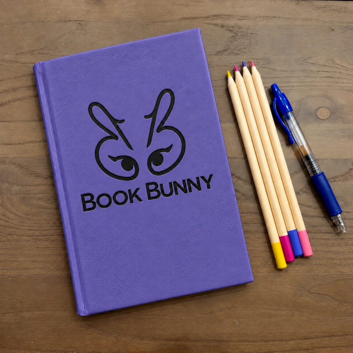 A purple hardcover notebook with a black bunny face logo and the text 'Book Bunny' on the cover, placed on a wooden tabletop next to four wooden pencils and a blue pen.
