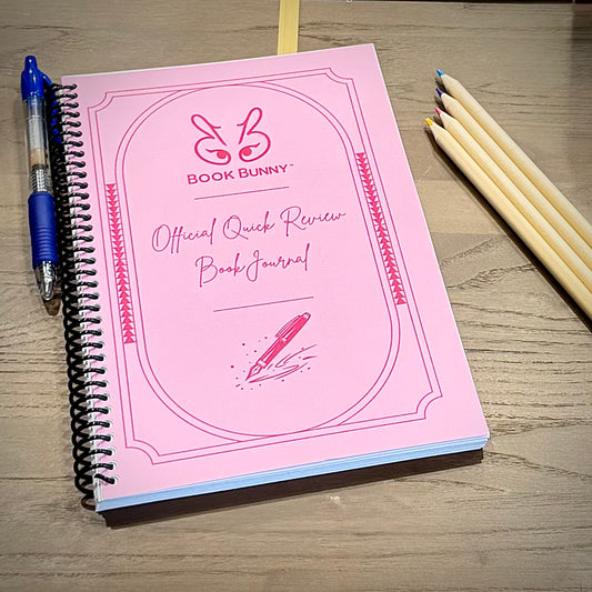 Pink Book Bunny™ Official Quick Review Book Journal with coil binding, shown with a blue pen and colored pencils on a wood surface.