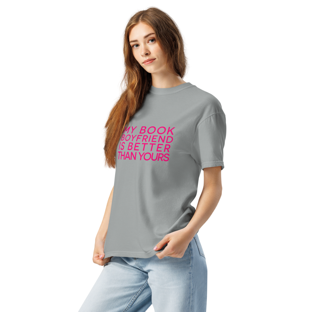 A woman modeling a gray crewneck t-shirt with the text 'MY BOOK BOYFRIEND IS BETTER THAN YOURS' printed in bright pink on the front, paired with light blue jeans against a plain white background.